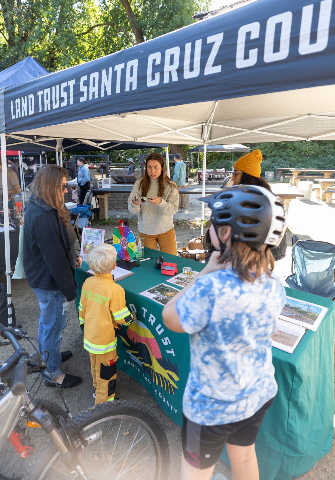 Children and adults at the Land Trust of Santa Cruz County booth under a blue canopy, looking at maps and displays.
